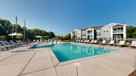 Located at 13511 Montoy Way in Charlotte, North Carolina, this outdoor amenity space centers around a generously sized resort-style swimming pool with crystal-clear turquoise water. The pool deck is finished in smooth concrete and lined with an abundance of white lounge chairs, offering ample space for sunbathing and relaxation under the Carolina sky. A shallow tanning ledge with in-pool loungers provides a distinctive recreational feature, allowing residents to recline directly at the water's edge.

The pool area is enclosed by a sleek black metal fence, providing both security and a clean aesthetic boundary. Strategically placed shade umbrellas offer relief from the sun, while manicured landscaping with young trees and ornamental plantings adds greenery throughout the deck perimeter.

Adjacent to the pool, a well-designed clubhouse building with board-and-batten siding in a soft teal-blue hue features a covered porch and large windows, suggesting interior amenity or social spaces. The surrounding residential buildings display a modern architectural style with gray and white siding, pitched rooflines, and private balconies. Mature trees frame the backdrop, providing natural privacy and a lush, park-like atmosphere that complements the overall community setting.
