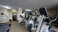 This fitness room offers a functional workout space designed to support residents' exercise routines. The room is outfitted with multiple cardio machines, including elliptical trainers and stationary bikes from recognized brands, arranged to allow comfortable use of each piece of equipment. Mirrored walls line one side of the space, visually expanding the room and allowing users to monitor their form during workouts. The flooring features a durable, textured surface well-suited for a fitness environment, providing both grip and resilience underfoot. Overhead fluorescent lighting illuminates the space evenly, ensuring good visibility throughout. A textured ceiling adds a subtle architectural detail overhead. Toward the back of the room, additional equipment is visible, suggesting a modest selection of strength or supplementary training options. A windowed entry door allows a degree of natural light to filter in while maintaining a sense of enclosure and privacy. The overall layout is practical and efficiently organized, making good use of the available space to deliver a convenient, on-site fitness option.