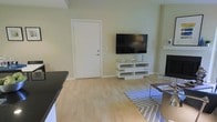 This single-floor space presents a well-organized living area characterized by light hardwood flooring that extends throughout, contributing to a bright and airy atmosphere. The room is anchored by a wall-mounted flat-screen television positioned above a modern white open-shelf media console, which provides practical storage while maintaining a minimalist aesthetic. To the right, a fireplace with a classic white mantle adds a traditional architectural element and serves as a secondary focal point within the space. The walls are finished in a soft, neutral tone that complements the light flooring and enhances the sense of openness. A patterned area rug in blue and grey tones defines the seating area and introduces a subtle decorative element. The room benefits from a straightforward, functional layout with a solid white interior door providing access to an adjoining area. The combination of clean lines, neutral finishes, and practical built-in features creates a cohesive and comfortable living environment suited for everyday use.