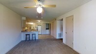 This single-floor residence offers an open-concept layout that seamlessly connects the living area and kitchen, creating a functional and cohesive living environment. The living space features wall-to-wall carpeting that adds warmth underfoot, while a ceiling fan with a light fixture provides both comfort and illumination overhead. A textured ceiling adds a classic architectural detail throughout the space.

The kitchen is positioned adjacent to the living area and is defined by a raised breakfast bar counter with two bar stools, making it an ideal spot for casual dining or conversation. The kitchen is equipped with dark wood cabinetry, a white refrigerator, and a built-in stovetop. Under-cabinet lighting brightens the workspace, and the countertop extends to form the convenient bar-height peninsula.

Beyond the main living and kitchen area, the layout opens into a hallway that leads to additional rooms, with multiple doors visible — suggesting separate bedroom and storage spaces. A floor-level HVAC vent is integrated into the wall, indicating a central climate control system. The neutral color palette of beige and white tones throughout lends the space a clean, understated appearance that is easy to personalize.