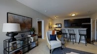 Located at 7505 Worthington Galena Rd in Columbus, Ohio, this single-floor residence offers a thoughtfully designed open-concept layout that seamlessly connects the living, dining, and kitchen areas. The living room is warm and inviting, featuring plush carpeting, neutral-toned walls, and stylish décor that creates a comfortable and modern atmosphere. A statement armchair with nailhead trim anchors the seating area, complemented by a decorative shelving unit and bold artwork that adds character to the space. The kitchen is outfitted with sleek dark cabinetry, granite-style countertops, and a breakfast bar with bar seating, making it both functional and visually striking. Track lighting overhead highlights the open flow between the kitchen and living areas. The dining area sits just beyond, offering an intimate setting for meals. A hallway visible from the main living area leads to additional private rooms, suggesting a well-organized floor plan. Throughout the space, the design strikes a balance between contemporary style and everyday livability, with cohesive finishes and purposeful details that make this residence a comfortable and attractive place to call home.