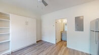 This single-floor space offers a thoughtfully designed layout with modern finishes and a cohesive aesthetic throughout. The kitchen is a functional highlight, featuring two-tone cabinetry — white upper cabinets paired with grey lower cabinets — granite-style countertops, a stainless steel sink, and a full suite of stainless steel appliances including a refrigerator, electric range, and over-the-range microwave. An open wood shelf adds a practical touch above the countertop.

The main living area is bright and airy, benefiting from vaulted ceilings that lend a sense of openness. A ceiling fan provides year-round comfort, while the warm-toned wood-look flooring runs continuously throughout the space, creating a seamless and inviting atmosphere. A large white built-in wardrobe unit with louvered doors and integrated shelving offers generous storage.

The bathroom is well-appointed with a grey vanity, granite-style countertop, a large mirror, and a walk-in shower with white tile surround and chrome fixtures. Wood-look flooring extends into the bathroom, maintaining the design continuity. The overall palette of whites, greys, and warm wood tones gives the entire space a clean, modern, and cohesive character.