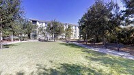 This multi-story residential building is situated within a well-maintained outdoor setting that emphasizes open space and natural surroundings. The building's exterior presents a clean, modern facade with a light neutral color palette, featuring rows of windows and balconies that offer residents outdoor access and views of the surrounding landscape.

The grounds are a defining feature of this property, with an expansive open lawn area providing generous green space. Mature trees, including tall pines and broad-canopied oaks, are distributed throughout the grounds, offering shade and a natural buffer between the building and its surroundings. Mulched planting beds add structured landscaping detail, while paved walkways wind through the property, connecting various areas of the grounds.

A fenced perimeter is visible along the building's frontage, suggesting dedicated amenity or recreational zones. The overall ambiance of the exterior is open, airy, and green, with the combination of maintained turf, mature tree canopy, and clear sightlines creating a park-like atmosphere. Under bright, clear skies, the property conveys a sense of calm and well-organized residential living within a natural setting.