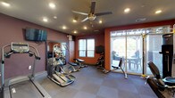 This fitness center is a thoughtfully designed, single-floor facility offering a comprehensive range of exercise options across multiple interconnected areas. The main cardio zone features several elliptical trainers and treadmills by brands such as Cybex and Precor, complemented by wall-mounted flat-screen TVs to keep members entertained during workouts. A ceiling fan overhead provides additional air circulation throughout the space. The strength training area is appointed with Hoist machines, a multi-purpose cable system, and a dedicated free weights section housing a full rack of dumbbells and an adjustable weight bench. Large mirrors line the walls to assist with form and technique. A separate functional training room provides ample open floor space and is stocked with stability balls, medicine balls, and organizational storage cubbies — ideal for stretching, core work, or group exercises. The facility is finished with durable, checkered carpet tile flooring throughout and is illuminated by recessed lighting. Natural light filters in through blinds-covered windows, while a set of sliding glass doors opens directly onto an outdoor covered patio furnished with seating, offering a pleasant view of the surrounding greenery. The bold accent walls in deep red and earthy tones lend the space an energetic and motivating ambiance.