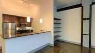 This single-floor space showcases a contemporary, open-concept design with an industrial aesthetic. The kitchen area is anchored by a sleek breakfast bar with dark granite countertops, providing both a functional workspace and a casual dining surface. Stainless steel appliances, including a microwave and range, are complemented by warm wood cabinetry, creating a balanced contrast of materials. Two cylindrical pendant lights hang above the bar, casting a warm, focused glow, while a track lighting system overhead ensures broader illumination throughout the space. Polished concrete floors run continuously throughout, contributing to the industrial character of the unit while remaining easy to maintain. A standout feature is the built-in open shelving unit positioned adjacent to the kitchen, consisting of multiple dark wood floating shelves supported by a structural beam overhead. These shelves offer generous display and storage capacity without enclosing the space. The overall palette is neutral, with light gray walls and warm wood tones creating a cohesive, modern atmosphere. The open layout and thoughtful design details make this a well-appointed, functional space suited to contemporary urban living.