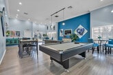Located at 1401 E Santo Antonio Dr in Colton, California, this single-floor space offers a thoughtfully designed open layout that seamlessly connects the living room, dining areas, and kitchen. The living area features light wood-style flooring, soaring ceilings with crown molding, and bold teal accent walls adorned with contemporary abstract artwork, creating a vibrant yet welcoming ambiance. Large windows fitted with white wood blinds allow ample natural light to fill the space while offering views of the surrounding exterior. The dining area accommodates multiple seating arrangements, including a round dark-topped table with upholstered blue chairs and a separate bar-height dining set, both bathed in natural light from generous window placements. A pendant light fixture adds a refined touch overhead. The kitchen is equipped with white cabinetry, dark countertops, stainless steel appliances including a full-size refrigerator, and a large window that brightens the workspace. Additional areas visible within the floor plan suggest dedicated workspaces or multipurpose rooms. A pool table anchors one section of the open layout, hinting at a community or recreational purpose. Throughout, the consistent use of light flooring, curated greenery, and cohesive art creates a polished, welcoming environment.