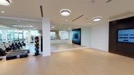 This contemporary fitness studio is designed with both functionality and aesthetics in mind, offering a bright and clean environment ideal for a wide range of workout activities. The space features light-toned hardwood-style flooring throughout, contributing to an open and welcoming atmosphere. Full-length, wall-to-wall mirrors line multiple walls, visually expanding the space while allowing users to monitor their form during exercise.

The studio is divided into distinct functional zones. A dedicated cycling area houses a row of stationary spin bikes, positioned near glass partitions that allow natural light to filter through from adjacent areas. A second zone is equipped for group fitness and functional training, featuring medicine ball racks, stability balls in multiple sizes, step platforms, and open floor space suited for stretching or mat-based workouts. A large flat-screen display mounted on a dark accent wall serves as a focal point for instructor-led programming.

Overhead, a series of modern flush-mount and drum-style ceiling light fixtures provide consistent, even illumination throughout the studio. The white walls and ceiling, combined with subtle wood-paneled accents visible through the glass partitions, lend the space a polished, upscale character. The open layout and thoughtful organization of equipment make this fitness studio a versatile and professional-grade training environment.
