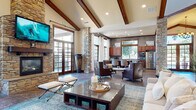 This well-appointed single-floor community clubhouse blends rustic charm with modern comfort through a cohesive design language of natural stone, rich wood tones, and contemporary furnishings. The main gathering area commands attention with soaring vaulted ceilings accented by prominent exposed wood beams and recessed lighting. A floor-to-ceiling stacked stone fireplace serves as the room's focal point, topped by a wall-mounted flat-screen television and flanked by French doors that invite abundant natural light and views of the outdoor area. Stone columns anchor the open floor plan, lending architectural character throughout. The lounge area offers generous seating arrangements on tile flooring with area rugs, creating a warm and inviting atmosphere. A fully equipped kitchen features rich cabinetry, granite countertops, a gas cooktop island with bar seating, stainless steel appliances, and mosaic tile backsplash — well-suited for events and gatherings. A separate media and lounge room includes patterned carpet, a mounted television, and sliding barn doors that open to a private workstation alcove with built-in granite desks and cabinetry. An entry corridor with tile flooring and stone column details provides a refined transition into the space. Together, these areas deliver a versatile, upscale environment designed for both relaxation and productivity.