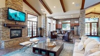 This well-appointed single-floor community clubhouse blends rustic charm with modern comfort through a cohesive design language of natural stone, rich wood tones, and contemporary furnishings. The main gathering area commands attention with soaring vaulted ceilings accented by prominent exposed wood beams and recessed lighting. A floor-to-ceiling stacked stone fireplace serves as the room's focal point, topped by a wall-mounted flat-screen television and flanked by French doors that invite abundant natural light and views of the outdoor area. Stone columns anchor the open floor plan, lending architectural character throughout. The lounge area offers generous seating arrangements on tile flooring with area rugs, creating a warm and inviting atmosphere. A fully equipped kitchen features rich cabinetry, granite countertops, a gas cooktop island with bar seating, stainless steel appliances, and mosaic tile backsplash — well-suited for events and gatherings. A separate media and lounge room includes patterned carpet, a mounted television, and sliding barn doors that open to a private workstation alcove with built-in granite desks and cabinetry. An entry corridor with tile flooring and stone column details provides a refined transition into the space. Together, these areas deliver a versatile, upscale environment designed for both relaxation and productivity.