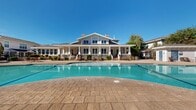 This inviting community space centers around a standout clubhouse building that showcases classic craftsman-inspired architecture, complete with a covered wraparound porch, white column accents, multi-pane windows, and charming roofline details. The exterior presents a clean, welcoming facade that overlooks the expansive outdoor amenity area below.

The focal point of the outdoor space is a generously sized, freeform swimming pool with striking blue tile accents and a shallow wading area, ideal for relaxation. The pool deck is finished with decorative stamped concrete pavers, providing ample room for lounge seating and outdoor furniture arranged throughout the space. A pergola-style cabana structure sits poolside, offering shaded gathering space for residents and guests.

Surrounding the pool, the grounds are thoughtfully landscaped with mature trees, tall cypress accents, and manicured shrubs that provide both privacy and aesthetic appeal. A wrought iron perimeter fence encloses the area, creating a secure and defined outdoor retreat. Scattered seating areas with tables and chairs, including umbrella-shaded dining spots, complete the social atmosphere of the space. Under clear, sunny skies, the entire outdoor setting exudes a resort-like ambiance designed for comfort and community enjoyment.