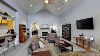 Located in Frisco, this single-story space impresses with its dramatic vaulted ceilings that soar to a sharp peak, lending an expansive, airy quality to the interior. The main living area is anchored by a classic fireplace with a decorative wood mantel, serving as a natural focal point for the room. Soft blue-gray walls complement the neutral carpeting, creating a calm and welcoming atmosphere throughout.

Two symmetrical arched entryways with elegant column detailing frame the transitions into adjacent areas, adding a refined architectural character to the space. These arches lead to what appear to be dedicated work or study nooks, each furnished with a desk and visible shelving, making the space highly functional.

A large bookcase filled with books and media lines one wall, reinforcing a sense of organized, purposeful living. Recessed lighting and a ceiling fan with an integrated light fixture provide both practical illumination and comfort. The living area is furnished with plush, earth-toned sofas and a sturdy wooden coffee table, offering ample seating for gathering or relaxing. A wall-mounted flat-screen television adds a modern touch. The overall design balances classic architectural details with everyday comfort and practicality.