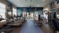 The fitness center at Haven at Bellaire, located in Richmond, TX, is a well-appointed, single-floor facility designed to support a comprehensive workout experience. The space is organized into distinct functional zones, each tailored to a specific type of training. The main cardio area features a row of Pro Maxima treadmills and elliptical machines positioned along large, light-filled windows with white plantation shutters, allowing ample natural light to energize the space. Oversized ceiling fans provide airflow throughout. A dedicated strength training zone houses a full rack of dumbbells, weight benches, and multi-use cable machines, with motivational wall graphics — including phrases like "Never Give Up," "No Excuses," and "No Pain No Gain" — adding an energetic atmosphere. A separate spin room offers stationary cycling bikes in a more intimate setting, with its own large window and dark accent wall. The zones are connected by elegant French doors with glass pane detailing, creating an open yet organized flow between areas. Dark rubber flooring and blue area runners run throughout, contrasting with the navy and neutral-toned walls that carry the Haven at Bellaire branding. Strategically placed mirrors expand the visual depth of the space, making it feel open and dynamic. The overall design is sleek, modern, and purpose-driven.