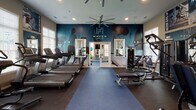 The fitness center at Haven at Bellaire, located in Richmond, TX, is a well-appointed, single-floor facility designed to support a comprehensive workout experience. The space is organized into distinct functional zones, each tailored to a specific type of training. The main cardio area features a row of Pro Maxima treadmills and elliptical machines positioned along large, light-filled windows with white plantation shutters, allowing ample natural light to energize the space. Oversized ceiling fans provide airflow throughout. A dedicated strength training zone houses a full rack of dumbbells, weight benches, and multi-use cable machines, with motivational wall graphics — including phrases like "Never Give Up," "No Excuses," and "No Pain No Gain" — adding an energetic atmosphere. A separate spin room offers stationary cycling bikes in a more intimate setting, with its own large window and dark accent wall. The zones are connected by elegant French doors with glass pane detailing, creating an open yet organized flow between areas. Dark rubber flooring and blue area runners run throughout, contrasting with the navy and neutral-toned walls that carry the Haven at Bellaire branding. Strategically placed mirrors expand the visual depth of the space, making it feel open and dynamic. The overall design is sleek, modern, and purpose-driven.