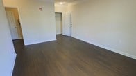 This single-floor space presents a clean and functional design that balances simplicity with comfort. The interior is finished with rich, dark hardwood flooring that runs throughout, providing a warm and grounding aesthetic against the neutral, light-toned walls. Wide white baseboards frame the perimeter of the room, adding a classic architectural detail that complements the overall clean aesthetic.

The main room is generously proportioned, offering a spacious and open feel with ample room for a variety of furniture arrangements. A built-in closet is conveniently situated near the entryway, providing practical storage with visible hanging rod space — an essential and well-integrated feature of the layout. A six-panel white door serves as the primary entrance, adding a traditional touch to the interior design.

Overhead lighting is provided via a flush-mount ceiling fixture, keeping the ceiling uncluttered and the space feeling open. Crown molding accents the top of the walls, lending a refined finish to what is otherwise a minimalist interior. The overall ambiance is calm, neutral, and move-in ready, offering a versatile blank canvas that can easily accommodate a range of interior styles and uses.