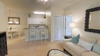 This single-floor residence offers a welcoming and cohesive open-plan layout, where the living area and kitchen flow naturally into one another. The living space is finished in soft, warm neutral tones and features plush carpeted flooring, creating a comfortable and inviting atmosphere. A ceiling fan overhead contributes to the airy feel of the room, while a stylish table lamp and accent furniture in cream and teal add a tasteful decorative touch.

The kitchen is well-appointed with crisp white cabinetry, white appliances, and a dark-topped breakfast bar that serves as a practical divider between the cooking and living areas. Two elegantly designed white bar stools with decorative circular back details are positioned at the bar, making it an ideal spot for casual dining or conversation. Upper cabinets extend to provide generous storage, and a plant on the countertop adds a touch of greenery.

Adjacent to the kitchen, a set of bifold doors leads to a closet, offering practical storage solutions. A bright exterior door allows natural light to filter into the space, enhancing the overall sense of openness. The warm color palette, consistent flooring, and thoughtful layout combine to create a harmonious and functional living environment.