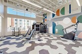 Located at 9910 W Montebello Ave in Phoenix, AZ, this fitness center is a thoughtfully designed exercise space that combines functionality with striking visual appeal. The room is defined by soaring ceilings with an exposed structural framework, flooded with abundant natural light through expansive floor-to-ceiling windows that offer views of the surrounding outdoor amenity areas. A standout feature is the large-scale, contemporary graphic mural that spans an entire wall, incorporating bold geometric shapes in teal, orange, green, and navy, adding a vibrant and energetic atmosphere to the space. The flooring features a distinctive hexagonal patterned carpet in neutral tones of grey and white, contributing to the modern aesthetic throughout. The fitness center is comprehensively equipped for a full-body workout experience, offering cardio machines including treadmills, ellipticals, a stair climber, and stationary bikes, as well as strength-training equipment such as a multi-use cable machine, adjustable benches, and a tiered dumbbell rack with a full range of free weights. Linear LED lighting complements the natural light, ensuring the space is well-illuminated at all times. A glass-panel door provides a visual connection to adjacent amenity spaces, reinforcing the open and welcoming character of this fitness facility.