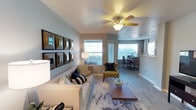 Located at 12200 Academy Rd NE in Albuquerque, New Mexico, this single-story residence offers a thoughtfully designed layout with comfortable living spaces and modern finishes. The open-concept living and dining area is bright and welcoming, featuring wood-look flooring, a ceiling fan with light fixture, and multiple windows that allow natural light to fill the space. The living room provides ample room for seating and entertaining, flowing seamlessly into the dining area, which is highlighted by a large window with outdoor views and elegant drapery. The kitchen is equipped with light wood cabinetry, black appliances including an electric range and built-in microwave, a double sink, and a breakfast bar that connects to the main living area. Each bedroom features plush carpeting, a ceiling fan, and a walk-in closet, offering generous storage and a restful atmosphere. The primary bedroom is particularly spacious with room for a large bed and additional furnishings. The bathrooms are well-appointed, featuring wide vanity mirrors, bright Hollywood-style lighting, and a combination tub and shower. Light wood cabinetry in the bathrooms complements the overall neutral and cohesive design palette found throughout the home. The textured ceilings and soft color tones contribute to a warm, comfortable ambiance.