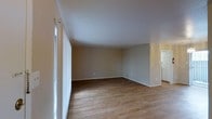 This single-floor space offers a clean, functional layout with a focus on simplicity and comfort. The main living area is generously sized, featuring warm hardwood-style flooring that runs throughout, lending a cohesive and inviting feel to the space. Neutral-toned walls in a soft beige palette provide a versatile backdrop, making the area easy to personalize with a variety of décor styles. Large windows along one wall allow natural light to filter in, brightening the interior and creating an airy atmosphere. The smooth white ceiling complements the light color scheme, enhancing the sense of openness. A doorway visible toward the back of the space provides access to an adjacent room, suggesting a practical and connected floor plan. The overall design is straightforward and uncluttered, offering a blank canvas well-suited for a range of uses. The combination of natural light, durable flooring, and neutral finishes gives this single-level space a move-in-ready quality that appeals to a wide range of occupants.