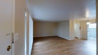 This single-floor space offers a clean, functional layout with a focus on simplicity and comfort. The main living area is generously sized, featuring warm hardwood-style flooring that runs throughout, lending a cohesive and inviting feel to the space. Neutral-toned walls in a soft beige palette provide a versatile backdrop, making the area easy to personalize with a variety of décor styles. Large windows along one wall allow natural light to filter in, brightening the interior and creating an airy atmosphere. The smooth white ceiling complements the light color scheme, enhancing the sense of openness. A doorway visible toward the back of the space provides access to an adjacent room, suggesting a practical and connected floor plan. The overall design is straightforward and uncluttered, offering a blank canvas well-suited for a range of uses. The combination of natural light, durable flooring, and neutral finishes gives this single-level space a move-in-ready quality that appeals to a wide range of occupants.