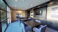 This contemporary lounge and dining space is thoughtfully designed with a refined, modern aesthetic. The room features multiple dark wood tables paired with plush, upholstered chairs in a muted blue-grey tone, creating a comfortable and stylish setting for dining or casual gatherings. Along one wall, floor-to-ceiling dark cabinetry with a matte finish provides ample storage, while a built-in counter with a subtly illuminated stone surface adds an elegant focal point. A wall-mounted flat-screen television is centrally positioned within the cabinetry, making the space well-suited for entertainment. On the opposite wall, a large-scale abstract artwork is displayed against a warm textured panel, adding a bold artistic element and visual contrast to the cool color palette of the room. Recessed lighting throughout the ceiling ensures even, ambient illumination, complementing the light blue carpeting that runs underfoot. The overall ambiance is polished and inviting, balancing functional design with contemporary style — ideal for communal dining, relaxation, or social interaction.
