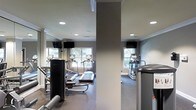 This fitness center is a thoughtfully designed, single-floor facility built to support a comprehensive workout experience. The space is outfitted with a variety of cardio equipment, including treadmills and elliptical machines, as well as strength training apparatus positioned throughout the room. Large windows line the walls, allowing natural light to fill the space and providing views of the surrounding greenery, creating an inviting and energizing atmosphere. Wall-to-wall mirrors are strategically placed to enhance the sense of openness and allow users to monitor their form during exercise. Mounted flat-screen televisions are positioned for easy viewing from the cardio machines, adding an entertainment element to workouts. Recessed ceiling lighting supplements the natural light, ensuring the space remains bright and well-lit at all times. The flooring is finished with a durable, cushioned surface appropriate for fitness use, while the neutral gray and white color palette on the walls contributes to a clean, modern aesthetic. Structural columns divide the space subtly without disrupting the overall flow. Crown molding adds a refined architectural touch, elevating the overall design above a standard gym environment.