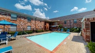 Located at 4010 Linkwood Dr in Houston's 77025 corridor, this single-story community facility serves as the welcoming centerpiece of the property. The entrance lobby immediately impresses with soaring double-height ceilings, large arched transom windows, and rich wood-framed double doors that flood the space with natural light. Elegant travertine tile flooring extends throughout the lobby, complemented by a striking tiered chandelier that anchors the room with a refined, upscale aesthetic. The open layout of the lobby provides clear sightlines to adjacent spaces, including a glass-partitioned area that houses a fitness center, visible through floor-to-ceiling windows fitted with horizontal blinds. Multiple wood-and-glass French doors provide seamless transitions between the interior and the outdoor amenity areas. Outside, the property showcases a generously sized rectangular swimming pool with a classic brick surround, finished in vibrant turquoise water. The pool deck is furnished with lounge chairs and a shaded patio table, ideal for relaxation. The surrounding brick exterior architecture ties the indoor and outdoor elements together cohesively, with lush greenery enhancing the overall curb appeal. This facility is well-suited to serve as a welcoming, functional amenity and leasing hub for the community.