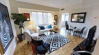 This single-floor residence offers a thoughtfully designed interior that balances classic charm with contemporary style. The living area serves as the heart of the space, showcasing warm parquet wood flooring arranged in a traditional pattern that adds visual depth and character throughout. Large windows dressed with horizontal blinds allow abundant natural light to fill the room, creating a bright and inviting atmosphere.

The living area is centered around a crisp white sofa and matching armchair, accented with teal throw pillows that introduce a vibrant pop of color. A bold black-and-white geometric area rug anchors the seating arrangement, complementing the glass-top coffee table and lending the space a modern edge. Framed artwork on the walls adds personality and warmth to the soft, neutral wall tones.

Adjacent to the main seating area, a compact dining space features a sleek black table and chairs, alongside a mirrored accent piece that enhances the sense of openness. The layout flows naturally between the living and dining zones, making the space feel connected and spacious. Recessed ceiling vents, elegant floor lamps, and carefully curated décor details throughout reflect a well-maintained and stylishly finished interior.