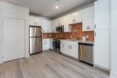 Located at 215 Willow Ave in Knoxville, Tennessee, this single-floor unit offers a clean, contemporary design with light wood-look flooring and neutral tones that flow seamlessly throughout the space. The open-concept kitchen is a standout feature, showcasing white shaker-style cabinetry with matte black hardware, stainless steel appliances including a range, microwave, dishwasher, and refrigerator, and a warm terracotta-tiled backsplash that adds a distinctive pop of color against the crisp white countertops. A utility closet with a utility hookup is conveniently accessible just off the kitchen area. The bedroom is bright and well-proportioned, featuring a ceiling fan, a large window that invites natural light and urban views of the surrounding Knoxville cityscape, and ample closet space. The bathroom is thoughtfully designed with a wide single-sink vanity topped with white counters, shaker-style cabinetry with matching black hardware, a large wall mirror with overhead lighting, and a tiled walk-in shower. Throughout the unit, recessed lighting, clean white baseboards, and a cohesive neutral palette contribute to a polished, move-in-ready aesthetic that suits modern urban living.