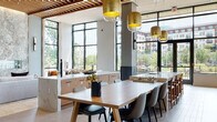 Located at 7970 Maitland St in McLean, Virginia, this single-floor amenity space delivers a polished, contemporary design that seamlessly blends communal dining, lounge, and social functions. The open-concept layout is anchored by soaring ceilings adorned with a striking slatted wood ceiling detail that adds warmth and visual texture throughout the central gathering area. Floor-to-ceiling black-framed windows wrap the perimeter, flooding the interior with abundant natural light and offering expansive views of the surrounding McLean streetscape and greenery. The kitchen and dining area features marble-surfaced islands with built-in seating, a large communal wood dining table with modern upholstered chairs, and gold-accented pendant lighting that adds a refined touch. Textured sage-green accent walls complement the neutral stone tile flooring found across the lounge zones. Multiple seating arrangements — from plush sectional sofas with colorful throw pillows to intimate two-person nook tables — provide versatile social spaces. A glazed door opens directly onto a covered outdoor terrace furnished with dining tables and string lights, extending the usable area outdoors. A reception desk anchors one end of the space, contributing to the welcoming, hospitality-inspired atmosphere of this well-appointed community hub.