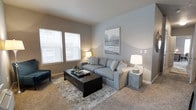 This single-floor unit is designed with an open-concept layout that seamlessly connects the living, kitchen, and dining areas into one cohesive and functional space. The living room is finished with wall-to-wall carpeting and neutral gray-toned walls, creating a calm and inviting atmosphere. Two large windows fitted with horizontal blinds allow natural light to fill the room, enhancing its airy feel. The space comfortably accommodates a sectional sofa, coffee table, and accent seating, making it well-suited for both relaxing and entertaining.

The kitchen is positioned at the heart of the unit, featuring warm wood-toned cabinetry, a black appliance suite, and a raised breakfast bar with seating for multiple barstools. The bar-height counter serves as a natural divider between the kitchen and the adjoining living area. A pendant light fixture highlights the dining area, which provides ample space for a full dining table and chairs. The flooring in the kitchen and dining zone transitions to a hard surface, offering a practical contrast to the carpeted living space. Overall, the unit presents a modern, neutral aesthetic throughout.