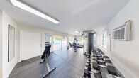 Located at 3600 Jackson St in Hollywood, Florida, this fitness center offers a single, well-appointed exercise room designed to support a comprehensive workout routine. The space features durable rubber interlocking floor mats covering the main workout area, complemented by tile flooring near the entrance, providing both functionality and easy maintenance.

The room is equipped with a variety of exercise equipment, including an elliptical machine, a cable weight machine, adjustable workout benches, and a multi-tiered dumbbell rack stocked with a range of hex weights. A full-length wall mirror allows users to monitor their form during exercise. Fitness reference charts are posted on the walls for guidance.

Natural light floods the space through large sliding glass doors and windows that line one wall, offering direct views of a sparkling outdoor pool surrounded by tropical palm trees — creating an energizing, resort-like atmosphere. A wall-mounted air conditioning unit ensures year-round comfort. Overhead fluorescent lighting and a skylight further brighten the interior.

The bright white walls and clean layout give the fitness center an open, airy feel. The sliding glass doors provide convenient direct access to the pool area, seamlessly connecting indoor fitness with outdoor recreation in this sunny South Florida setting.