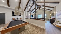 Located at 83 Terrace Greene Dr in Barboursville, VA, this single-floor community clubhouse offers a thoughtfully designed gathering space that blends rustic charm with modern sophistication. The room is immediately defined by its dramatic vaulted ceiling, adorned with dark-stained exposed wood beams and intersecting steel truss framework that draws the eye upward and creates an impressive sense of vertical volume.

The living area is anchored by a plush brown leather sectional sofa paired with sage green accent chairs, centered around a low coffee table atop a layered area rug. A textured slatted wood accent wall serves as a striking focal point, framing a wall-mounted television flanked by warm sconce lighting and complemented by a row of dark navy cabinetry below.

Large, floor-to-ceiling black-framed windows line the walls, flooding the space with natural light and offering views of the surrounding neighborhood. Light wood-look plank flooring runs throughout, contributing to the warm, inviting ambiance.

A dedicated game zone features a pool table beneath a sculptural woven pendant light fixture, alongside a shuffleboard table positioned beneath the soaring steel truss partition. Additional recreational options, including an arcade game, round out the entertainment offerings. The space is well-lit with recessed lighting and globe pendant fixtures, creating a balanced and welcoming atmosphere throughout.