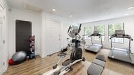 Located at 450 Waldan Cir in Acworth, Georgia, this fitness center is a compact yet thoughtfully appointed workout facility designed to support a well-rounded exercise routine. The space features hardwood-style flooring in the main workout area, transitioning to carpet in the cardio zone, and is finished with crisp white walls and a textured ceiling with recessed lighting throughout.

The cardio section is equipped with multiple treadmills, stationary spin bikes, and an elliptical trainer, all positioned to face windows that overlook lush, tree-lined greenery — providing a pleasant, nature-infused workout experience. A wall-mounted TV is also present for entertainment during exercise. A dedicated strength and functional training area houses a TAGfit medicine ball rack stocked with a variety of weighted balls, multiple stability and exercise balls, and a weight bench, offering ample options for strength, balance, and core training.

Large windows and glass-paneled doors allow generous amounts of natural light to fill the room, creating a bright and energizing atmosphere. Multiple entry points, including a standard door and a glass-paned door, contribute to easy accessibility. The overall design is clean, functional, and efficient, making effective use of the available space to deliver a comprehensive fitness experience.