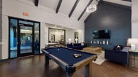 This single-floor community amenity space is designed with a contemporary aesthetic, blending industrial and modern elements throughout. The centerpiece of the space is a dramatic great room showcasing soaring vaulted ceilings with bold dark exposed beams, polished concrete floors, and a striking dark shiplap accent wall fitted with a wall-mounted flat-screen TV and built-in cabinetry. Two navy blue tufted sofas flank a solid wood coffee table, creating an inviting lounge area beneath an elegant chandelier. A full-size pool table anchors the open game area, which flows seamlessly into the fully equipped kitchen. The kitchen features professional-grade stainless steel appliances, including a commercial range with hood, double wall oven, and large refrigerator, complemented by white cabinetry, a soft blue subway tile backsplash, and a dark-topped communal dining island with pendant lighting overhead. A separate lounge area offers a large sectional sofa, wall-mounted TV, and curated black-and-white framed photography. A dedicated workspace includes a built-in desk, computer workstation, and additional seating. Black-framed glass doors and windows connect the interior to outdoor areas, allowing abundant natural light to fill the space throughout.