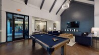 This single-floor community amenity space is designed with a contemporary aesthetic, blending industrial and modern elements throughout. The centerpiece of the space is a dramatic great room showcasing soaring vaulted ceilings with bold dark exposed beams, polished concrete floors, and a striking dark shiplap accent wall fitted with a wall-mounted flat-screen TV and built-in cabinetry. Two navy blue tufted sofas flank a solid wood coffee table, creating an inviting lounge area beneath an elegant chandelier. A full-size pool table anchors the open game area, which flows seamlessly into the fully equipped kitchen. The kitchen features professional-grade stainless steel appliances, including a commercial range with hood, double wall oven, and large refrigerator, complemented by white cabinetry, a soft blue subway tile backsplash, and a dark-topped communal dining island with pendant lighting overhead. A separate lounge area offers a large sectional sofa, wall-mounted TV, and curated black-and-white framed photography. A dedicated workspace includes a built-in desk, computer workstation, and additional seating. Black-framed glass doors and windows connect the interior to outdoor areas, allowing abundant natural light to fill the space throughout.