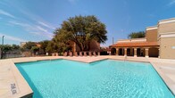 This outdoor pool area is defined by its generous scale and Mediterranean-inspired architectural surroundings. The centerpiece is a large, sparkling swimming pool with clearly marked depth zones, complemented by a separate spa/hot tub for relaxation. The expansive concrete sundeck surrounds the pool on multiple sides, providing ample space for the numerous lounge chairs positioned throughout the area.

The adjacent clubhouse building features distinctive Spanish-style architecture, characterized by terra cotta roof tiles, stucco exteriors, and a series of arched colonnades that frame a covered walkway along the poolside. This covered corridor runs the length of the building, offering shaded passage and direct pool views through its decorative arched openings and iron railings. Unit entrances along this colonnade are accessed via a dark paneled door with glass detailing.

The pool area is fully enclosed by an iron security fence with a gated entry, ensuring controlled access. Mature trees provide natural shade and visual interest throughout the space. The entire area benefits from abundant natural sunlight, creating a bright, resort-like atmosphere ideal for outdoor recreation.