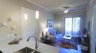 This single-floor residence offers a thoughtfully designed open-concept layout that seamlessly connects the kitchen and living areas. The kitchen is equipped with granite countertops, a stainless steel sink with a modern gooseneck faucet, and elegant pendant lighting that casts a warm glow over the workspace. Crown molding adds a refined architectural touch throughout the space, enhancing the overall sense of polish and attention to detail. The living area is bright and welcoming, centered around a comfortable sofa accented with decorative pillows and complemented by a stylish coffee table. A ceiling fan with integrated lighting ensures year-round comfort. Warm hardwood flooring runs throughout, adding richness and continuity to the space. Framed wall art contributes a personal, curated aesthetic to the room. A sliding glass door at the far end of the living area opens to a private balcony, allowing natural light to filter in and providing a pleasant outdoor retreat. The neutral color palette of soft grays and whites creates a calm, versatile backdrop that suits a variety of interior styles. The overall ambiance is modern, comfortable, and move-in ready.