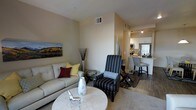 This single-floor residence offers a thoughtfully designed open-concept layout that connects the living area and kitchen in a cohesive, functional flow. The living room serves as the central gathering space, featuring warm neutral-toned walls, hard-surface flooring, and a soft area rug that anchors the seating arrangement. A light gray sofa with accent pillows and a patterned armchair provide comfortable seating around a round marble-top coffee table, while decorative elements such as tall vase arrangements and wall art add character to the space. The dining area sits adjacent to the living room, furnished with bar-height seating that bridges the transition into the kitchen. The kitchen is equipped with white cabinetry, stainless steel appliances including a refrigerator and microwave, and a well-lit countertop workspace illuminated by recessed lighting. The overall ambiance is bright and inviting, with a clean, contemporary aesthetic throughout. Consistent hard-surface flooring in the kitchen and dining zone contrasts with the soft carpeting in the living area, creating a natural visual distinction between the zones while maintaining an open, connected feel throughout the space.