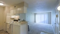 This single-floor space is designed with an open-concept layout that seamlessly connects the kitchen and living area. The kitchen features white cabinetry, a light-toned countertop with a built-in breakfast bar, and a white electric range with an overhead range hood. A soffit detail above the upper cabinets adds architectural definition to the cooking space, while a small bar stool at the counter provides a casual dining option. The kitchen opens directly into the living area, creating a cohesive and functional flow throughout the main living space. The living area is finished with plush, neutral-toned carpeting that adds warmth and comfort underfoot. A large floor-to-ceiling window fitted with vertical blinds serves as a prominent feature of the living space, allowing abundant natural light to fill the room and creating a bright, airy atmosphere. The walls are painted in a soft, neutral tone, complementing the light color palette maintained throughout. A textured ceiling adds subtle visual interest overhead. The overall ambiance of the space is clean, functional, and welcoming, with a cohesive design that maximizes the open floor plan.