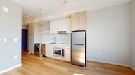 This contemporary single-floor unit showcases a clean, minimalist design with light hardwood flooring throughout, creating a cohesive and airy atmosphere across every room. The open-concept layout allows the kitchen and living area to flow seamlessly together, maximizing the sense of space.

The kitchen is efficiently designed along a single wall, featuring light wood and white cabinetry that extends to the ceiling for ample storage. Stainless steel appliances include a refrigerator, a built-in microwave oven, and a cooktop with an overhead range hood. A stainless steel sink with a modern faucet completes the workspace, while a white subway tile backsplash adds a clean, polished finish. In-unit laundry — a washer and dryer — is conveniently integrated directly beside the kitchen cabinetry.

The living area is bathed in natural light, courtesy of large windows that frame open outdoor views and allow sunlight to pour across the light wood floors. Soft, neutral gray walls complement the bright aesthetic throughout.

The bathroom maintains the same understated, modern character, with clean white walls, a vanity with storage drawers, and warm hardwood flooring. Track lighting installed on the ceilings provides functional illumination across the unit, reinforcing the space's practical yet contemporary design sensibility.