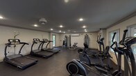 This fitness center offers a comprehensive workout environment designed to accommodate a wide range of exercise routines. The space features dark, durable rubber flooring throughout, providing both safety and functionality underfoot. Recessed lighting and ceiling fans ensure the area remains well-lit and comfortable during use.

Cardio equipment is prominently represented, with multiple treadmills, elliptical machines, a stationary bike, and a rowing machine distributed throughout the room. A dedicated strength-training section includes cable machines, a multi-function weight station, and a full rack of dumbbells organized along a mirrored wall, which also visually expands the space.

Natural light enters through several windows along the perimeter walls, offering views of the surrounding greenery. Multiple entry and exit doors provide convenient access. A wall-mounted climate control unit supports year-round comfort. Medicine balls and additional free-weight accessories round out the available equipment.

The neutral gray walls paired with the dark flooring create a clean, focused atmosphere. The layout allows ample open floor space between machines, supporting both individual workouts and ease of movement throughout the facility.