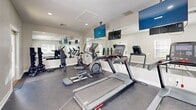 Located at 7959 Cottage Hill Rd in Mobile, Alabama, this dedicated exercise room offers a well-appointed fitness facility designed to support an active lifestyle. The single-room space is finished with a durable dark floor surface well-suited for high-traffic athletic use, while neutral-toned walls and crisp white baseboards and trim create a clean, motivating environment.

The fitness area is equipped with multiple cardio machines, including treadmills, an elliptical, and a stationary bike, providing a well-rounded selection of cardiovascular workout options. A full-length wall mirror spans one side of the room, visually expanding the space and allowing users to monitor their form during exercise. Mounted flat-screen televisions are positioned for easy viewing from the equipment, enhancing the workout experience.

Natural light filters in through several windows fitted with white horizontal blinds, offering views of the surrounding greenery and keeping the space feeling bright and open. Recessed ceiling lighting supplements the natural light, ensuring the room remains well-lit at any hour. A glass-paneled entry door provides convenient exterior access and further contributes to the airy atmosphere. Free weights are also available, rounding out the functional amenities of this efficiently designed fitness space.