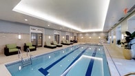 Located at 659 Rice Boulevard in Exton, Pennsylvania, this indoor aquatic facility offers a polished and well-appointed environment designed for both fitness and relaxation. The centerpiece of the space is a full-length lap pool with clearly marked swimming lanes, finished in crisp blue tile and illuminated by recessed underwater lighting that enhances the clarity of the water. The pool area is lined with stylish chaise lounges accented in green cushions, providing a comfortable space for rest and recovery. The surrounding deck is finished in light neutral tile, contributing to the clean, spa-like aesthetic of the room. Architectural details include a tray ceiling with soft cove lighting that casts a warm, even glow throughout the space, complemented by recessed overhead fixtures and wall-mounted sconces that maintain a bright yet inviting ambiance. French-style glass doors along one wall introduce natural light and add a classic design element to the interior. A hot tub or hydrotherapy feature is visible at the far end of the pool, adding a wellness component to the facility. The overall atmosphere is refined and tranquil, making this space equally suited for dedicated lap swimming and leisurely relaxation.