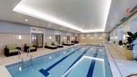 Located at 659 Rice Boulevard in Exton, Pennsylvania, this indoor aquatic facility offers a polished and well-appointed environment designed for both fitness and relaxation. The centerpiece of the space is a full-length lap pool with clearly marked swimming lanes, finished in crisp blue tile and illuminated by recessed underwater lighting that enhances the clarity of the water. The pool area is lined with stylish chaise lounges accented in green cushions, providing a comfortable space for rest and recovery. The surrounding deck is finished in light neutral tile, contributing to the clean, spa-like aesthetic of the room. Architectural details include a tray ceiling with soft cove lighting that casts a warm, even glow throughout the space, complemented by recessed overhead fixtures and wall-mounted sconces that maintain a bright yet inviting ambiance. French-style glass doors along one wall introduce natural light and add a classic design element to the interior. A hot tub or hydrotherapy feature is visible at the far end of the pool, adding a wellness component to the facility. The overall atmosphere is refined and tranquil, making this space equally suited for dedicated lap swimming and leisurely relaxation.