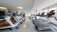 Located at 7601 River Rd in North Bergen, NJ, this contemporary fitness center offers a well-appointed, single-floor workout facility designed with both function and aesthetics in mind. The space is characterized by a striking curved ceiling detail with circular flush-mounted light panels that lend a modern, architectural feel to the interior. Large floor-to-ceiling windows line the perimeter walls, flooding the facility with abundant natural light and providing open views of the surrounding neighborhood streetscape.

The workout floor features durable, easy-to-maintain rubberized flooring in a cool grey tone, providing a safe and supportive surface throughout. The cardio equipment section is generously stocked with multiple treadmills and elliptical trainers, all positioned to take advantage of the expansive window views. Additional equipment includes stationary bikes, a rowing machine, and a dumbbell rack with free weights, ensuring a comprehensive range of workout options for users of varying fitness levels.

The open layout allows for comfortable circulation and ample spacing between machines. The overall ambiance is clean, bright, and modern, creating an inviting environment conducive to focused exercise. The combination of thoughtful architectural design, plentiful natural light, and a diverse array of fitness equipment makes this facility a well-rounded and functional amenity space.