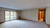 Located at 1798 Magnolia Ln N in Plymouth, Minnesota, this single-floor home offers a practical and comfortable layout designed for easy living. The expansive living room is a standout feature, with plush carpeting, warm wood baseboards, and a textured ceiling that adds character. A large sliding glass door floods the space with natural light and provides direct access to an outdoor area, offering pleasant views of the surrounding greenery. The open flow from the living room leads seamlessly into the dining and kitchen area. The kitchen is well-appointed with warm oak cabinetry, dark laminate countertops, light hardwood-style flooring, and white appliances including a range, over-the-range microwave, refrigerator, and dishwasher. A ceiling fan overhead keeps the space comfortable. The bathroom is clean and functional, featuring white tile flooring, an oak vanity with a single sink, a large mirror with overhead lighting, and a full tub-shower combination. Neutral tones throughout the home create a cohesive and inviting atmosphere. The layout, as seen in the dollhouse view, is thoughtfully organized, with clearly defined living, kitchen, and private spaces making this home both efficient and welcoming.