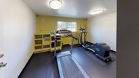 This fitness center is a thoughtfully organized exercise facility designed to support a well-rounded workout routine. The space is divided into distinct functional zones, each serving a specific fitness purpose. The main cardio area is outfitted with a treadmill, elliptical trainer, and upright stationary bike, all positioned on protective rubber floor mats and illuminated by flush-mount ceiling lights that provide consistent, even lighting throughout. A vibrant yellow accent wall adds an energizing pop of color to the cardio zone, which also features a window that invites in natural light and a shelving unit for storing accessories such as yoga mats and small equipment. An adjacent area serves as a dedicated strength and functional training zone, equipped with a rack of resistance bands, kettlebells of varying weights, and additional hand weights neatly arranged along the wall. A posted workout chart provides exercise guidance for users. A large wall mirror in this section visually expands the space and supports proper form during workouts. The open floor area offers ample room for stretching, bodyweight exercises, or floor-based routines. The dark flooring throughout ties the space together with a sleek, gym-appropriate aesthetic, and the overall layout ensures ease of movement between stations. Clean white and neutral walls keep the environment feeling fresh and focused.