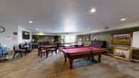 The Mountain View Community Room at 200 S Ironton St in Aurora, Colorado, is a well-appointed, single-floor gathering space designed to accommodate a variety of recreational and social activities. The room is generously sized, offering an open and comfortable layout throughout.

At the heart of the space sits a classic billiard table with a bold red felt surface, serving as a central focal point. Surrounding it are multiple dining and game tables paired with wooden chairs, ideal for board games, cards, or casual meals — a chess set and checkerboard are among the games available. An upright piano lines one of the deep olive-green accent walls, accompanied by framed artwork that adds a warm, decorative character to the room.

A dedicated lounge area features a plush leather sofa and armchairs arranged around a flat-screen TV, creating a relaxed viewing nook. Recessed lighting and natural light from windows along the perimeter keep the space bright and welcoming. Lush indoor plants are positioned throughout, adding a touch of greenery.

The adjacent kitchen area is fully functional, featuring warm wood cabinetry, dark granite countertops, a central island, a full-size range, refrigerator, and microwave — well-suited for hosting events and gatherings. The textured carpet flooring throughout ties the space together, contributing to its inviting, community-centered atmosphere.