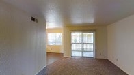 This single-story space features an open living area that benefits from abundant natural light, delivered through two key sources: a fixed window and a full-width sliding glass door that opens to an exterior balcony or patio. The sliding door is dressed with vertical blinds, offering flexible light and privacy control, while framing views of the surrounding outdoor environment.

The living area is finished with wall-to-wall carpeting, providing a warm and comfortable underfoot feel, while an adjoining zone transitions to hard flooring, creating a natural distinction between spaces. The textured popcorn ceiling adds a classic architectural detail overhead, complemented by a centrally positioned HVAC vent that ensures climate comfort throughout.

The walls are painted in a soft, neutral tone that enhances the brightness of the space and lends a clean, understated backdrop. The layout is straightforward and functional, with an open flow that connects the main living zone to the adjacent area. Overall, the space presents a practical and light-filled interior well-suited for comfortable everyday living.