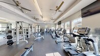 This fitness center offers a well-appointed, single-floor layout designed to accommodate a full range of workout routines. The space features a clean, modern aesthetic with crisp white walls, a tray ceiling with recessed lighting, and multiple ceiling fans that promote air circulation throughout. Large windows line the walls, flooding the interior with natural light and offering views of an outdoor pool area, creating an open and energizing atmosphere. The durable rubber flooring provides both safety and comfort underfoot, suited for high-traffic athletic use. A large wall-mounted mirror expands the sense of space while supporting form-focused training. The cardio section is well-stocked with treadmills, elliptical machines, stationary bikes, and stair climbers from True Fitness and similar brands, many equipped with individual display screens. The strength training area features a dedicated free weight section with a full dumbbell rack, multiple weight benches, and barbell stations with plate-loaded squat racks. Selectorized cable and resistance machines round out the strength equipment offerings. Wall-mounted flat-screen televisions are positioned throughout the space for entertainment during workouts. Diamond-plate metal wall accents add an industrial touch, complementing the overall contemporary design of this thoughtfully outfitted fitness facility.